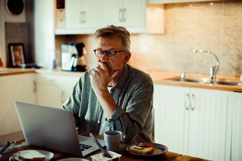 Man sitting at kitchen table reading his laptop.